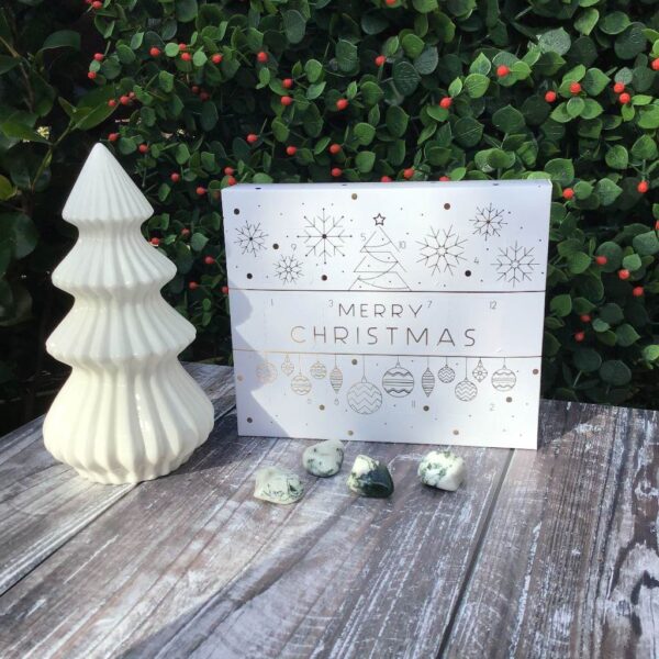 White advent calendar with gold lettering sitting alongside a white tree decoration and white and green tumblestone crystals.