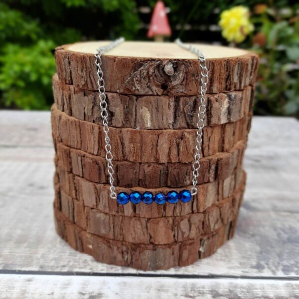 Handmade Blue Hematite Necklace. Features a row of faceted beads suspended from a stainless steel chain. Necklace draped over stack of wood slices.