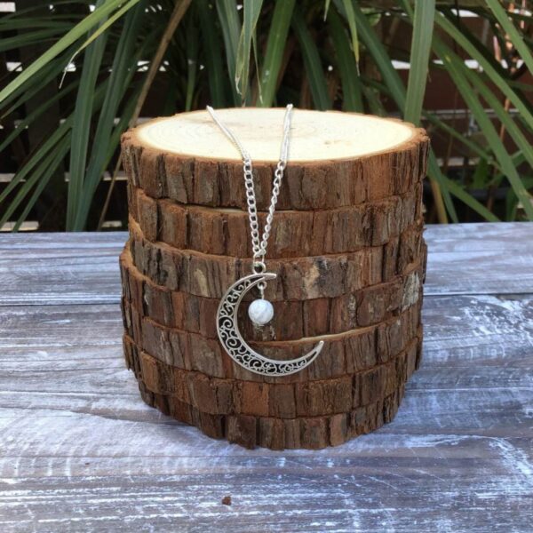 Handmade moon charm necklace. Made with stainless steel chain and silver tone charm with a white/grey Howlite bead. Necklace shown draped over stack of wood slices.